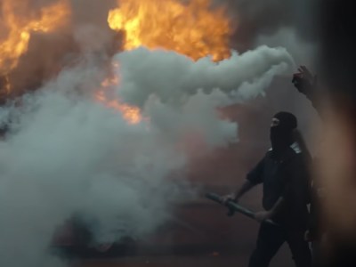 A black-clad rioter against a backdrop of smoke and flame from Suburraeterna.