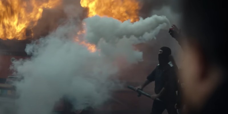 A black-clad rioter against a backdrop of smoke and flame from Suburraeterna.
