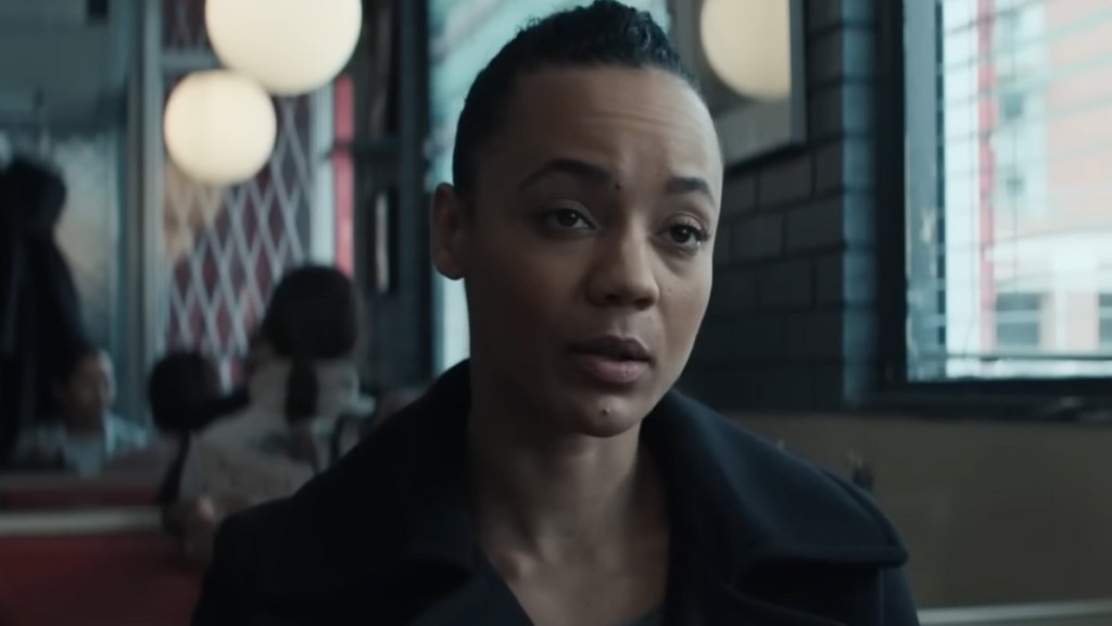 A short-haired woman in a black coat sitting in a diner in Reacher. 