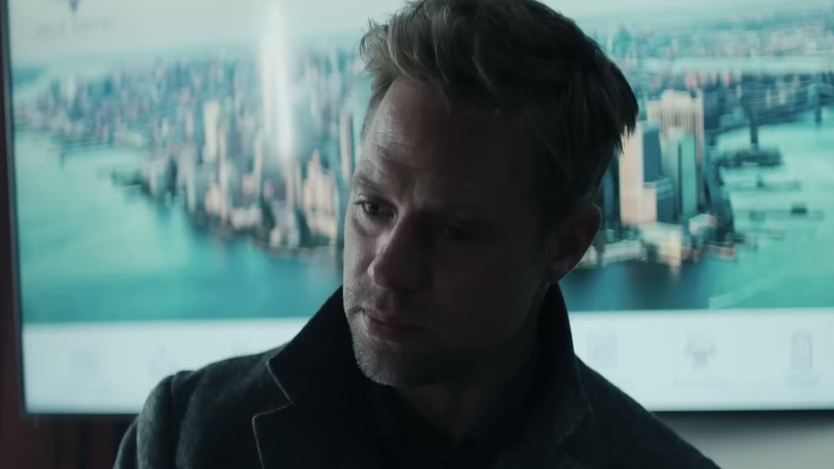 A rough-shaven man in a winter coat, standing in front of a photo of new York in Reacher. 