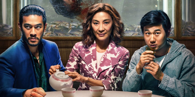 A woman and two younger men sitting at a table, drinking tea. A severed head is floating in a fish tank behind them. This image is part of an article about all the major actors and the cast list for The Brothers Sun.